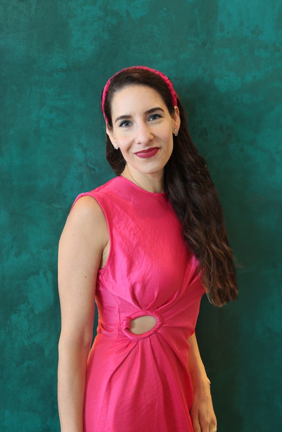 Latina woman with long hair held back by a pink headband gently smiling in a bright pink dress against a teal wall.